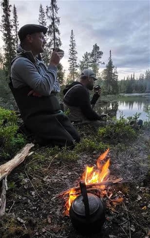 En grupp människor i vildmarken vid Jormvattnets fiskecamp, omgivna av skog och utrustade för camping. De sitter kring en lägereld, där en kastrull värms, och en person bär en hatt.