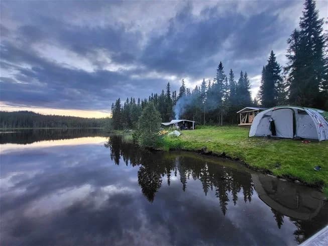 En stilla sjö omgiven av tät barrskog vid Jormvattnets fiskecamp, spegelblankt vatten med reflektioner av träd och en klar himmel ovanför.