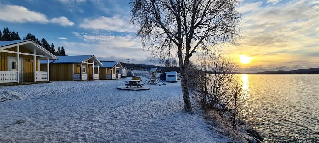 En vintervy över Jormvattnets Fiskecamp med en stuga vid sjökanten i skymningen, omgiven av frusna vattendrag och snöbeklädda marker.