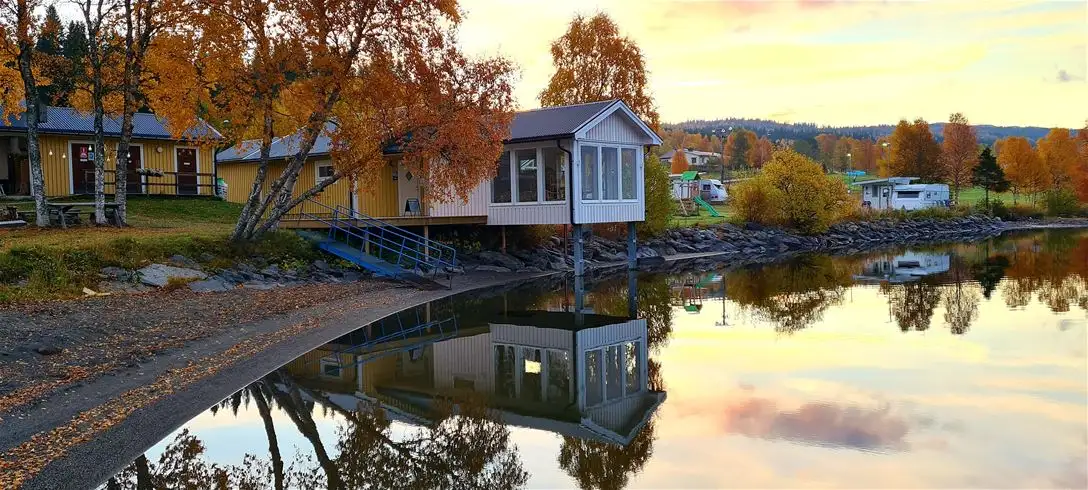 En naturskön bild av Jormvattnets Fiskecamp, där stugor speglas i det stilla vattnet av en sjö omgivet av frodig grönska.