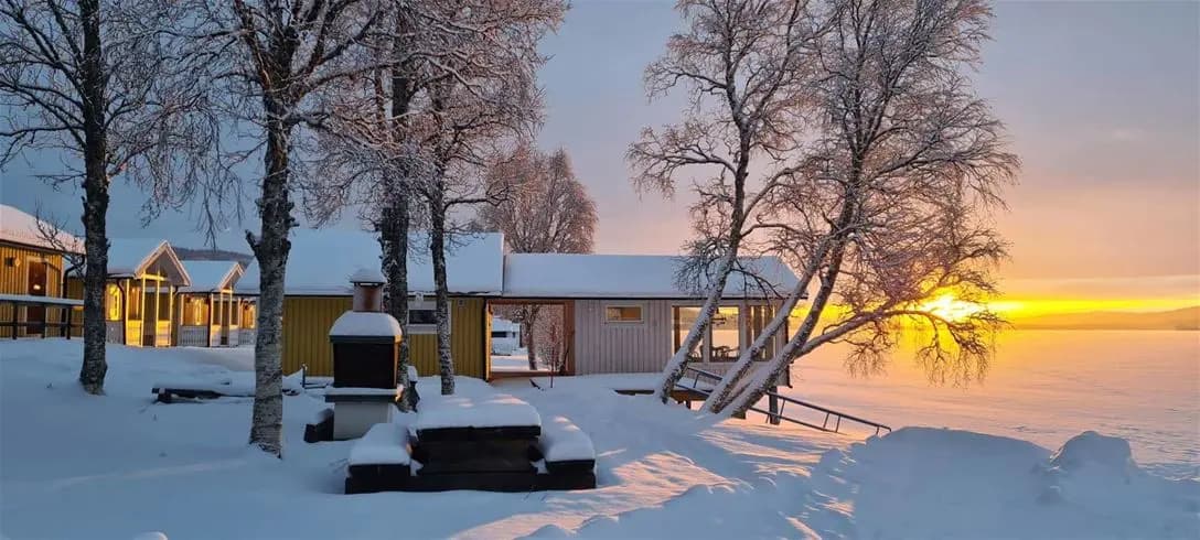 En snötäckt stuga vid Jormvattnets fiskecamp i skymningen, omgiven av vinterträd och en fridfull atmosfär.