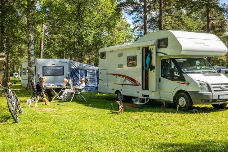 En semesterhusbil och husvagn är uppställda på älvdalens camping, omgiven av grönska och en lugn skogsmiljö.