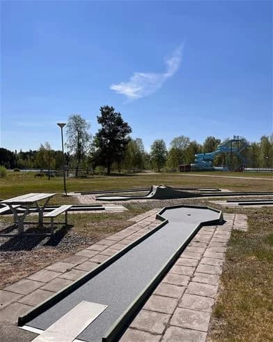 En grönskande park vid Hede Camping med en upptrampad gångväg som slingrar sig genom gräsmattan. I bakgrunden syns bänkar för avkoppling och utomhusvistelse.