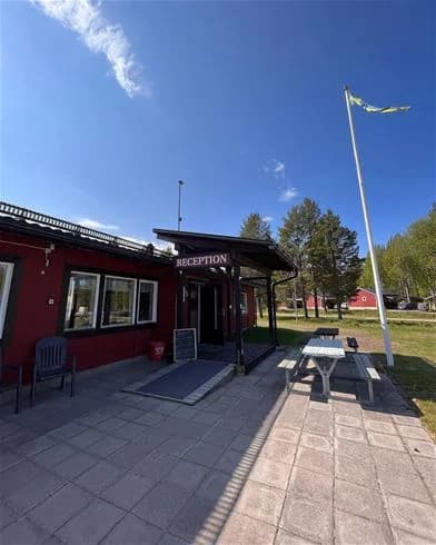 Bild av Hede Camping, med skuggade viloområden och utemöbler under ett solskydd. Pen och vilobänkar är utspridda på den välskötta gården, vilket skapar en inbjudande plats för besökare att koppla av. I bakgrunden syns byggnadssidor som hör till campingens faciliteter.