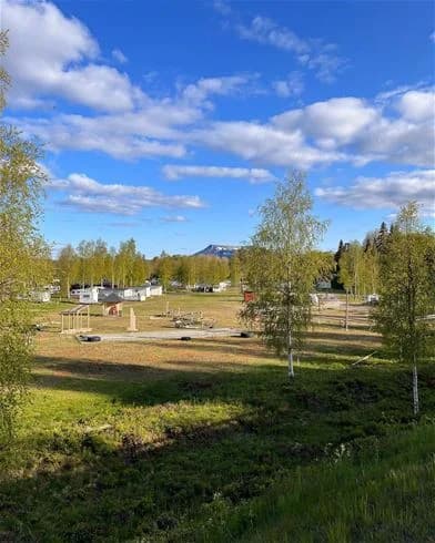Vy över Hede Camping med gräsmatta i förgrunden, omgiven av träd och natur. Himlen är delvis molntäckt.