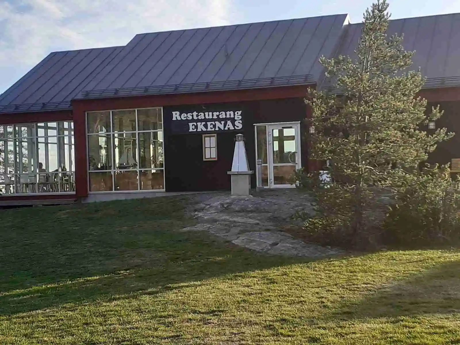 En lugn atmosfär vid Ekenäs gästhamn & camping, med utsikt över en restaurangdel. Bord och stolar står uppradade utomhus, inbjudande för besökare som vill njuta av en måltid eller fika i en naturnära miljö.