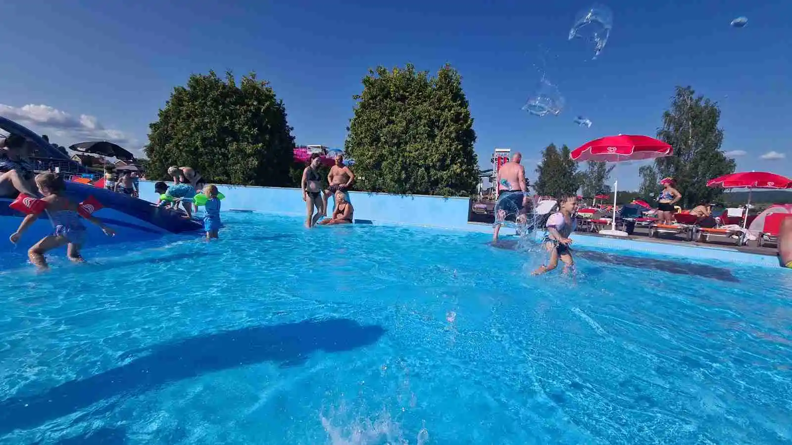 En solig dag vid Sunne Camping och Sommarland, där besökare njuter av sommarens aktiviteter. Folk i baddräkter leker och simmar i en stor utomhuspool, omgivna av glada semesterfirare.