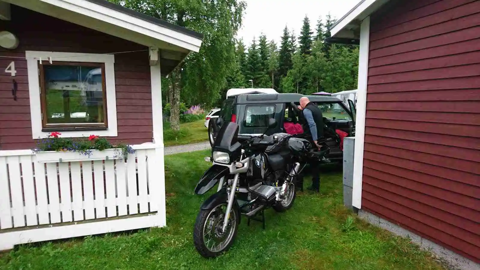 En campingplats med en motorcykel parkerad vid sidan av ett tält. Motorcykelns detaljer som hjul, däck och belysning är tydligt synliga. I bakgrunden skymtas träd och andra campingtillbehör.