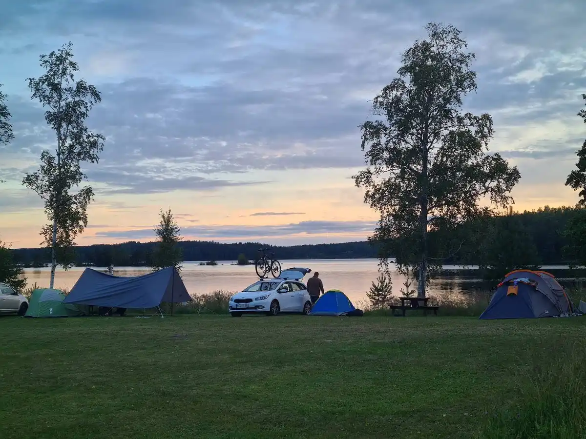 Bild av Uskavigårdens camping vid skymningen, där tält är uppsatta nära en sjö. Området är omgivet av grönska och tältplatserna skyddas delvis av presenningar. En fridfull atmosfär med inslag av rekreation i en naturskön miljö.
