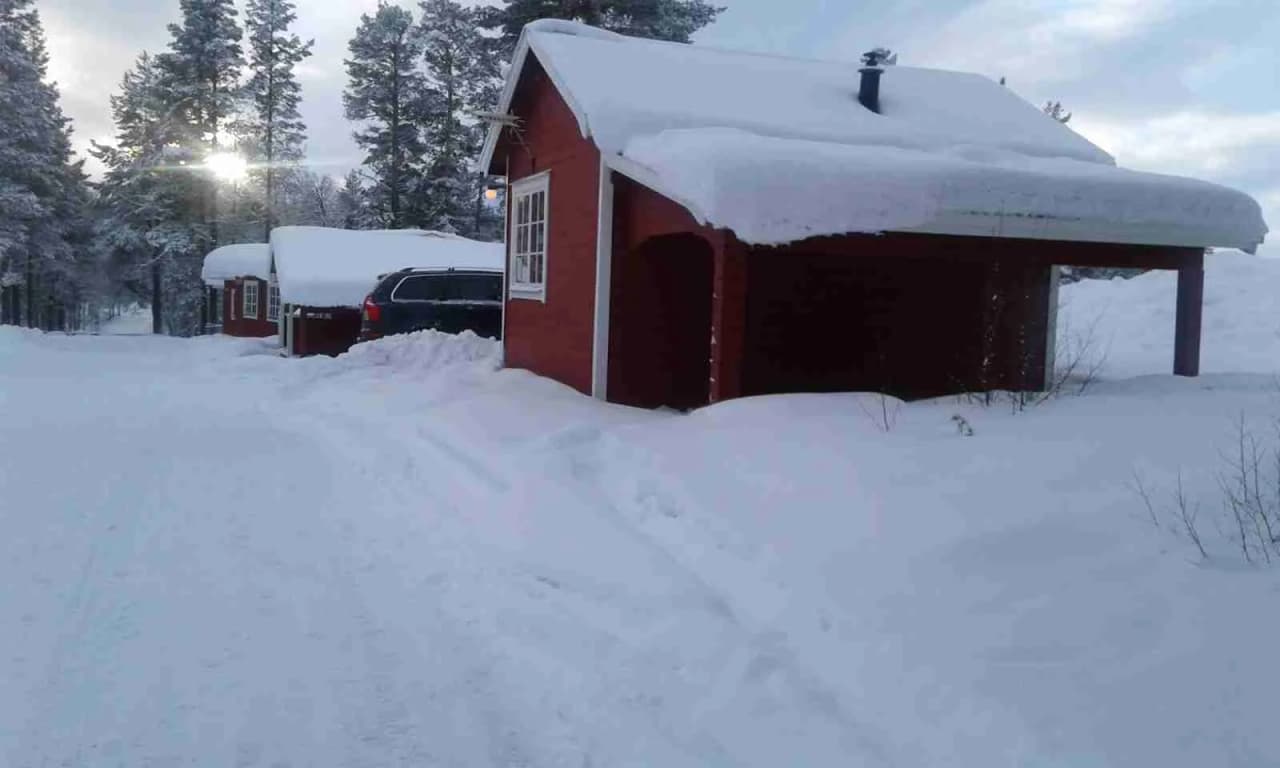 En snötäckt stuga på Vemdalens camping, omgiven av vinterlandskap med tungt snöbelagda tak och snötyngda träd i bakgrunden.