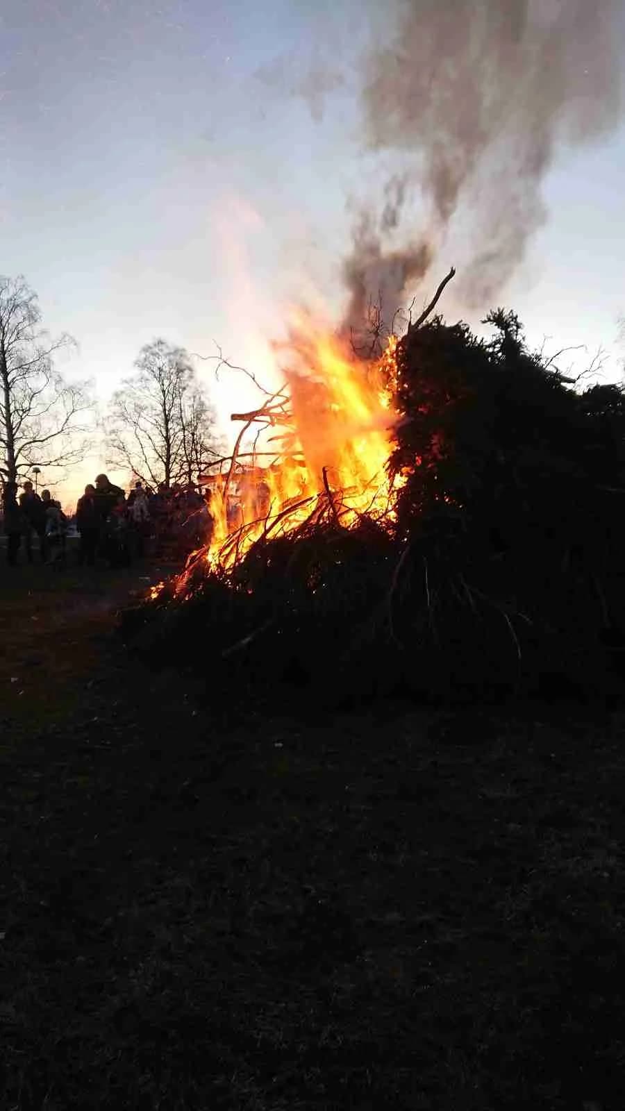 En stämningsfull brasa i skymningen med rök som stiger mot himlen, omgiven av öppna fält och en lätt vind.