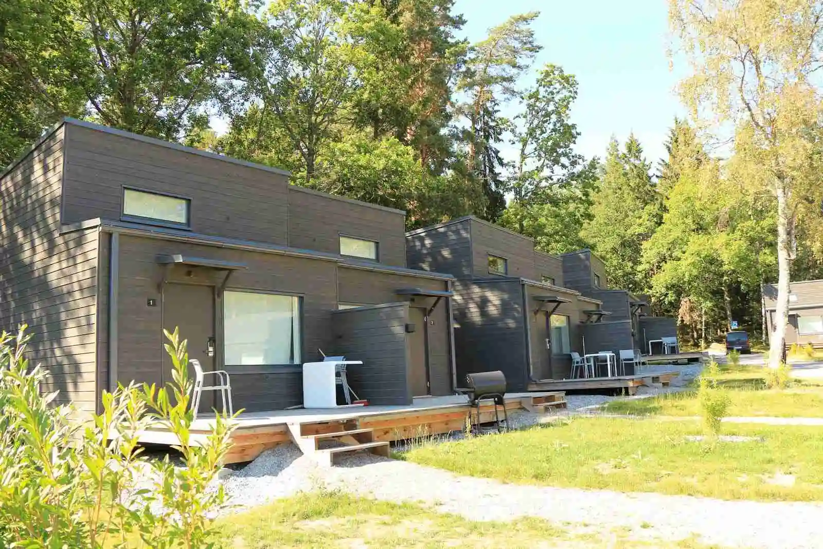 En hemtrevlig stuga omgiven av grönska på Rösjöbaden Camping & Stugby, med en välskött uppfart och stencirkulerad gångväg som leder genom trädgården. Evergreen-träd skapar en fridfull bakgrund.