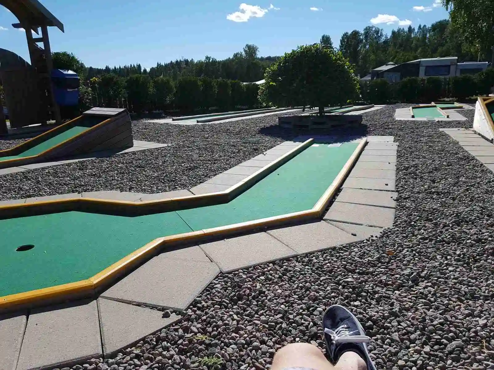 En bild av Charlottenberg camping & stugor - Haganäset som visar en minigolfbana omgiven av gröna gräsytor och landskapsarkitektur. Omgivningen är skuggad och har inslag av konstgräs, vilket skapar en inbjudande parkatmosfär.