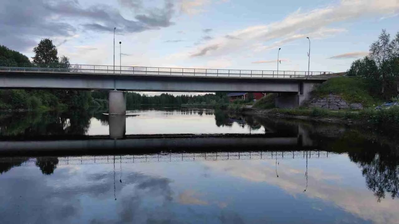 En bro sträcker sig över en bred vattenväg och speglas i dess yta. Vattnet omges av frodiga, gröna stränder.