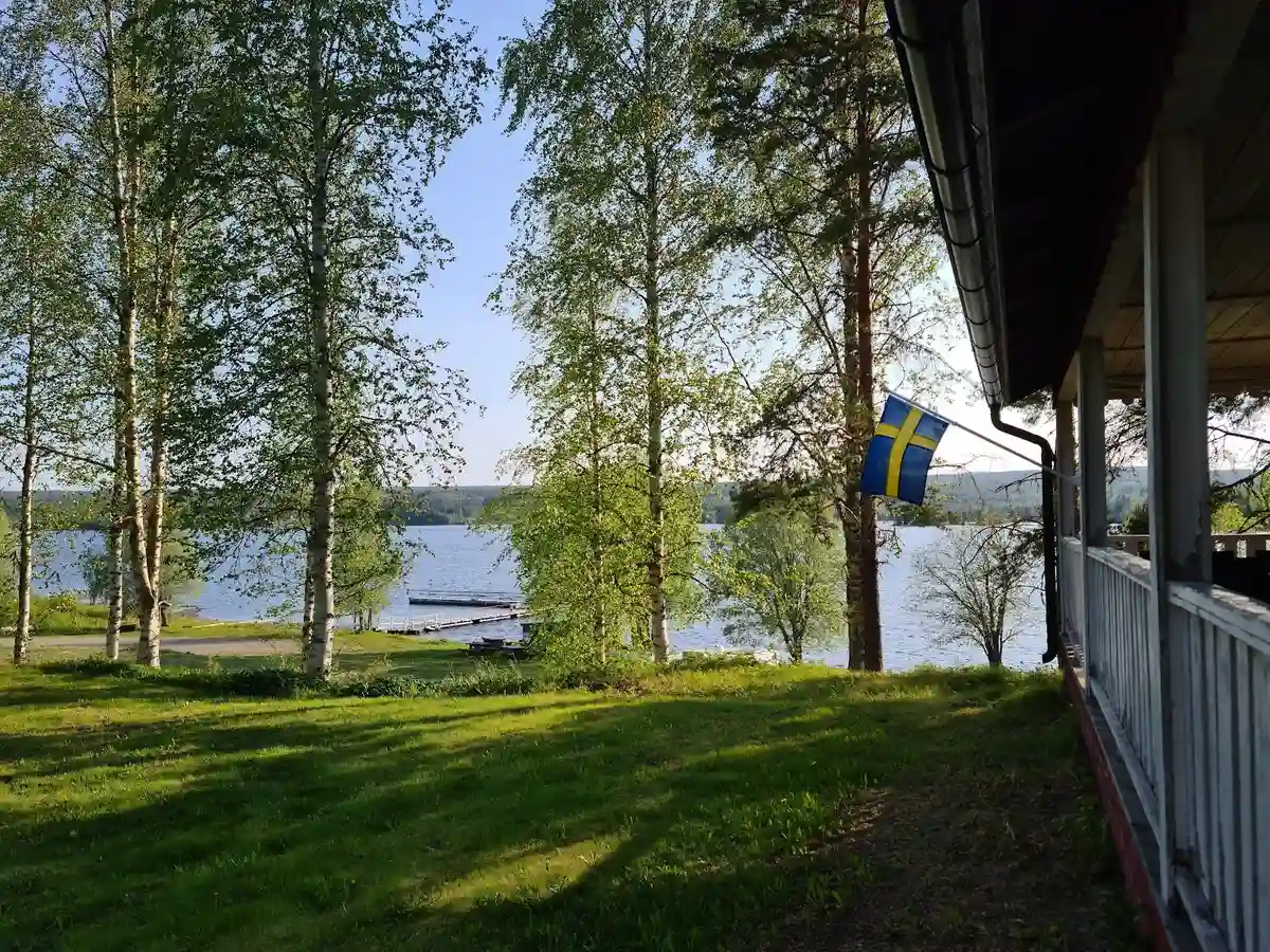 En grönskande campingplats med skuggiga områden och en välskött gräsmatta vid kanten av en sjö. I bakgrunden syns en kuperad landskap med träd och vattendrag, vilket skapar en fridfull och naturskön miljö.