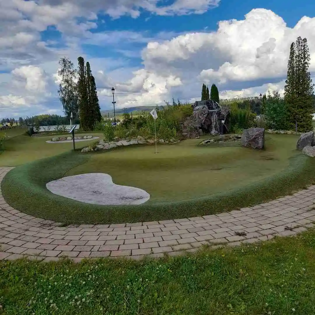 En grönskande parkmiljö på Sunne Camping och Sommarland med välskött gräsmatta, vackra landskapsdetaljer, minigolfbana, stigar och barrträd. En vattenanläggning syns i bakgrunden.