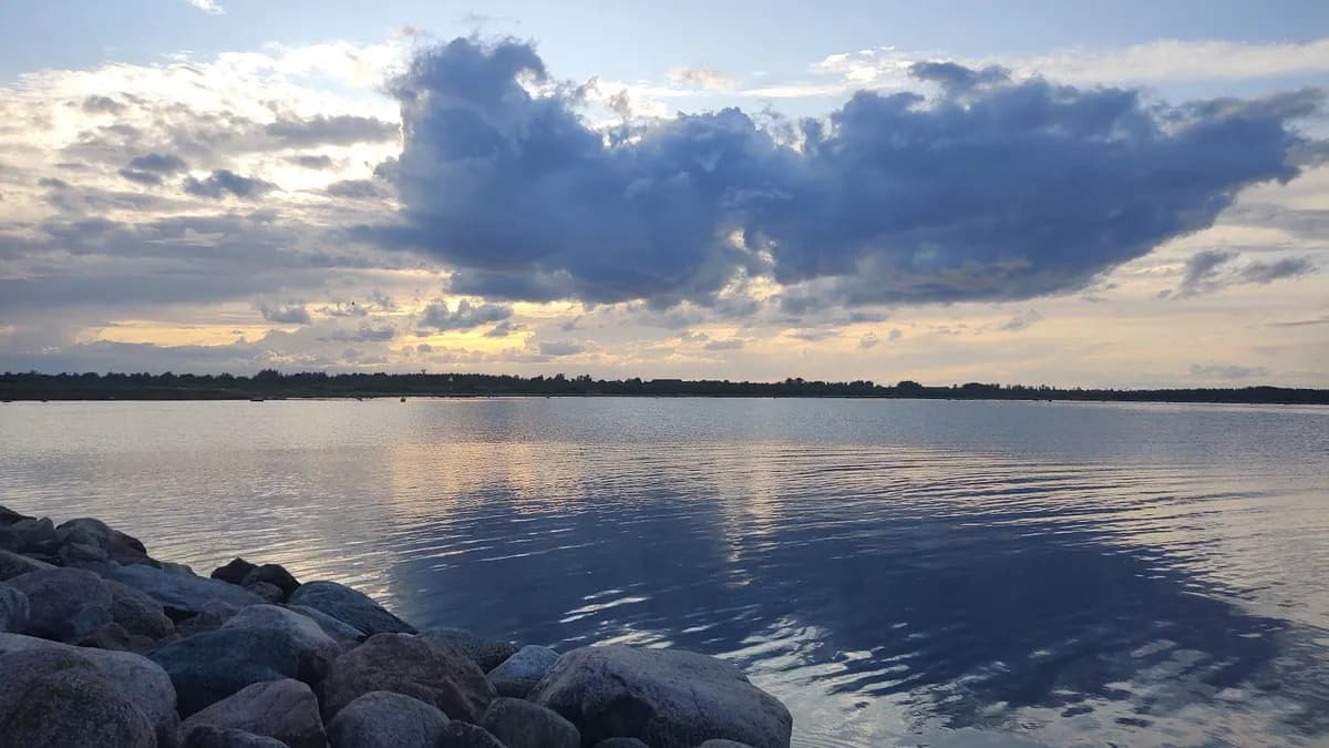 Bild av en somrig och klar dag vid Gröndals Camping & Stugor. Den blå himlen är delvis täckt av vita moln, och i bakgrunden syns en horisont över ett stilla vattendrag.