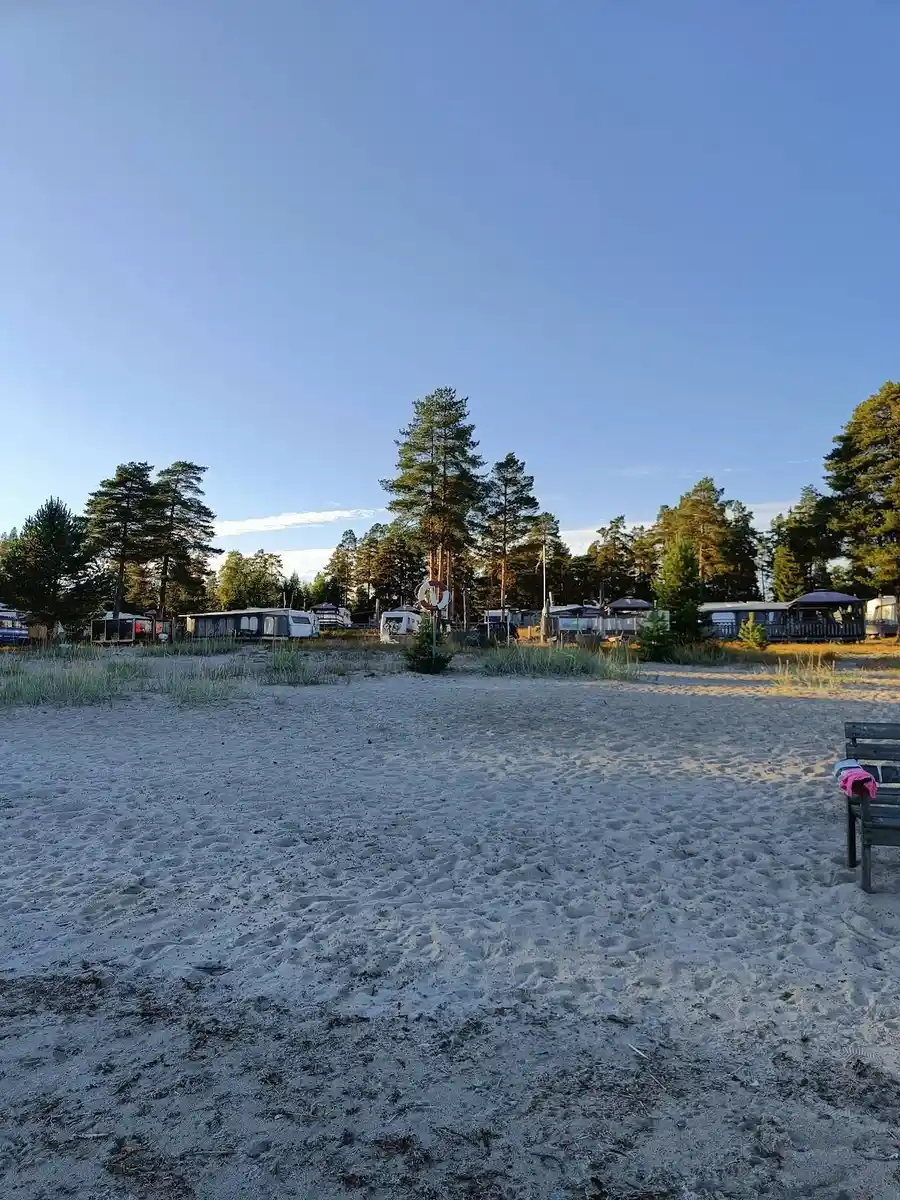 Bild av en solig sommardag vid Ava havsbad och camping. Sandstrand möter lugna vågor under en blå himmel med vita, fluffiga moln.