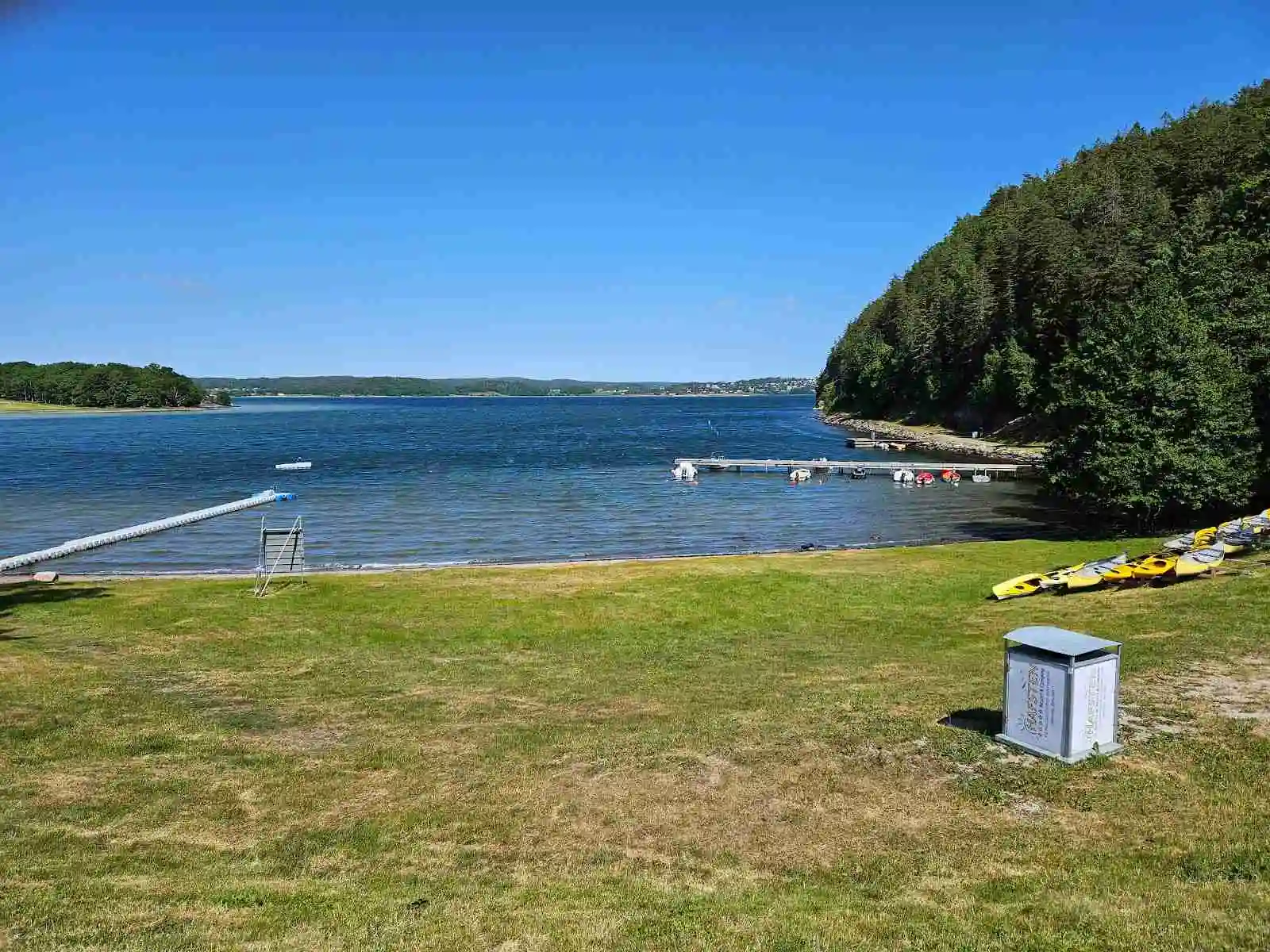 En lugn sjö vid Hafsten Resort & Camping, omgivande skog och kullar med en strandlinje. Vattenfarkoster syns på vattnet i bakgrunden.