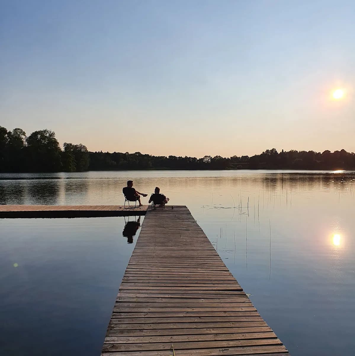 En stillsam sjö omgiven av grönska med en liten brygga som sträcker sig ut över vattnet. Speglingar av landskapet syns i sjöns yta.