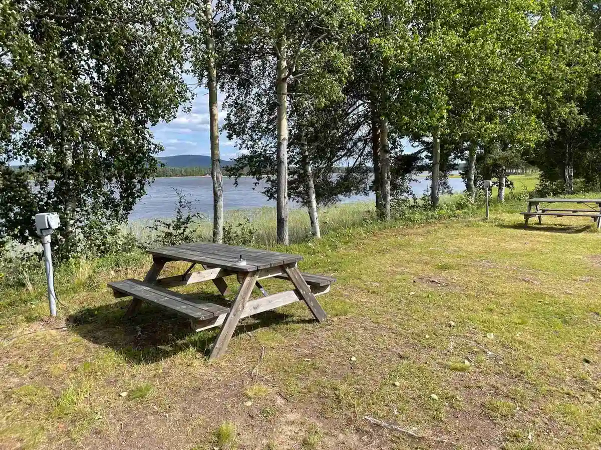 En picnicplats med bord och bänkar nära en sjökant, omgiven av grönskande växtlighet vid Trollforsen Camping & Cottages.