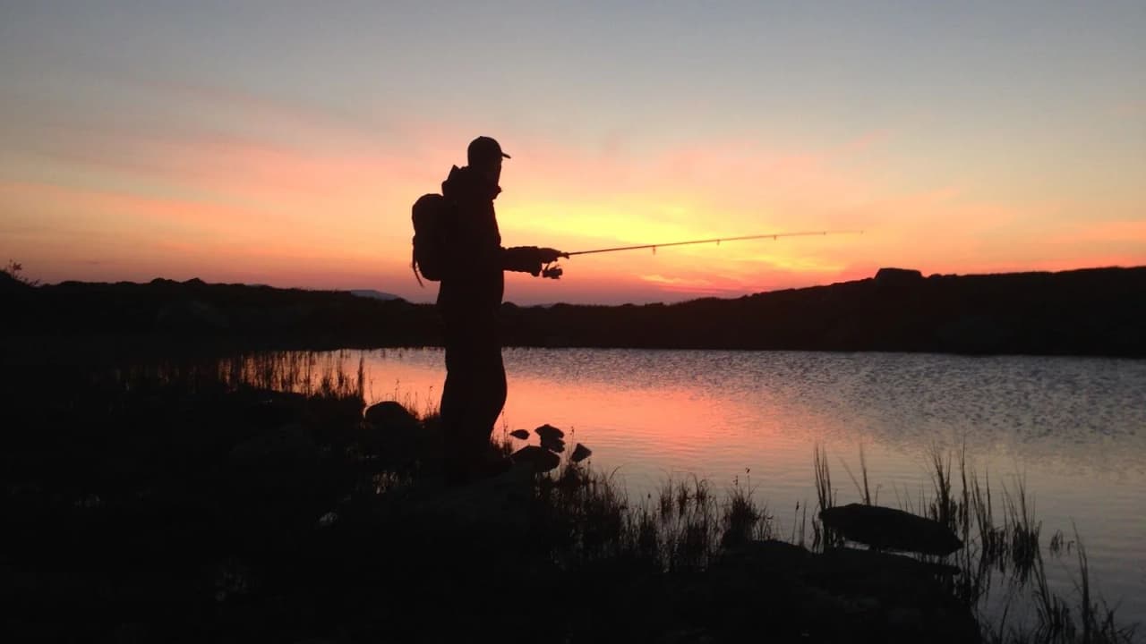 En solnedgång över Rörvattnets fiskecamp, där en fiskare med fiskespö står vid vattnet och njuter av kvällens rofyllda stämning.