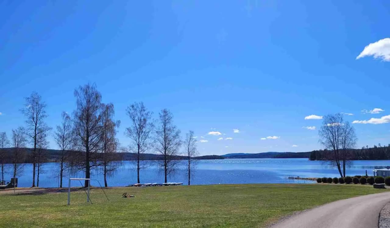 En vacker vy av en klarblå sjö vid Charlottenberg camping & stugor - Haganäset, med en horisont som möter den blå himlen. I förgrunden syns gröna kullar och träd som omger sjön, vilket skapar en fridfull och naturskön atmosfär.