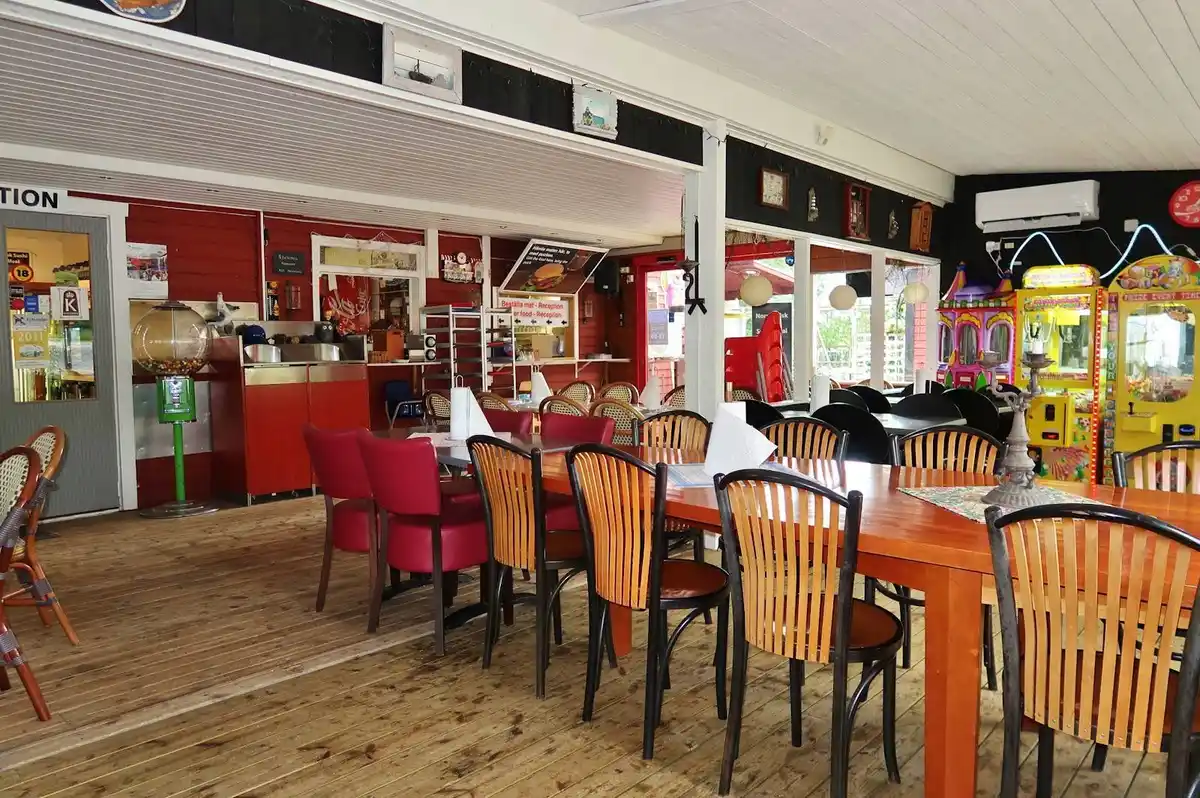 En utomhusrestaurang med träbord och stolar under sommarsäsong, perfekt för en måltid nära Ava havsbad och camping. Varm och inbjudande atmosfär med trädetaljer.