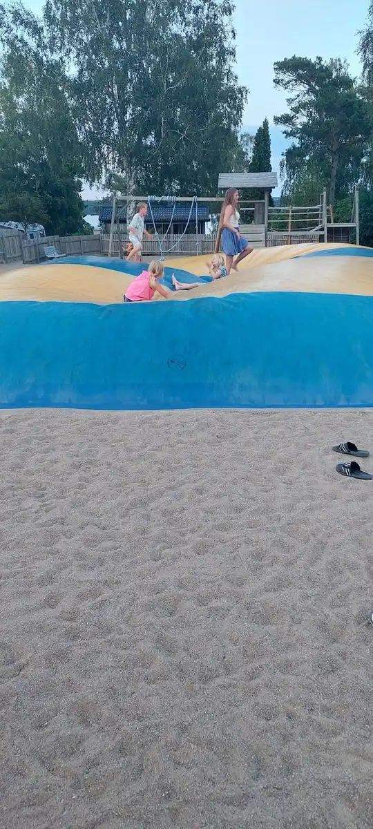 Bild av en lekplats vid Almöns bad & camping, med sand, rutschkana och lekområde omgiven av träd som ger skugga, perfekt för rekreation och semester.