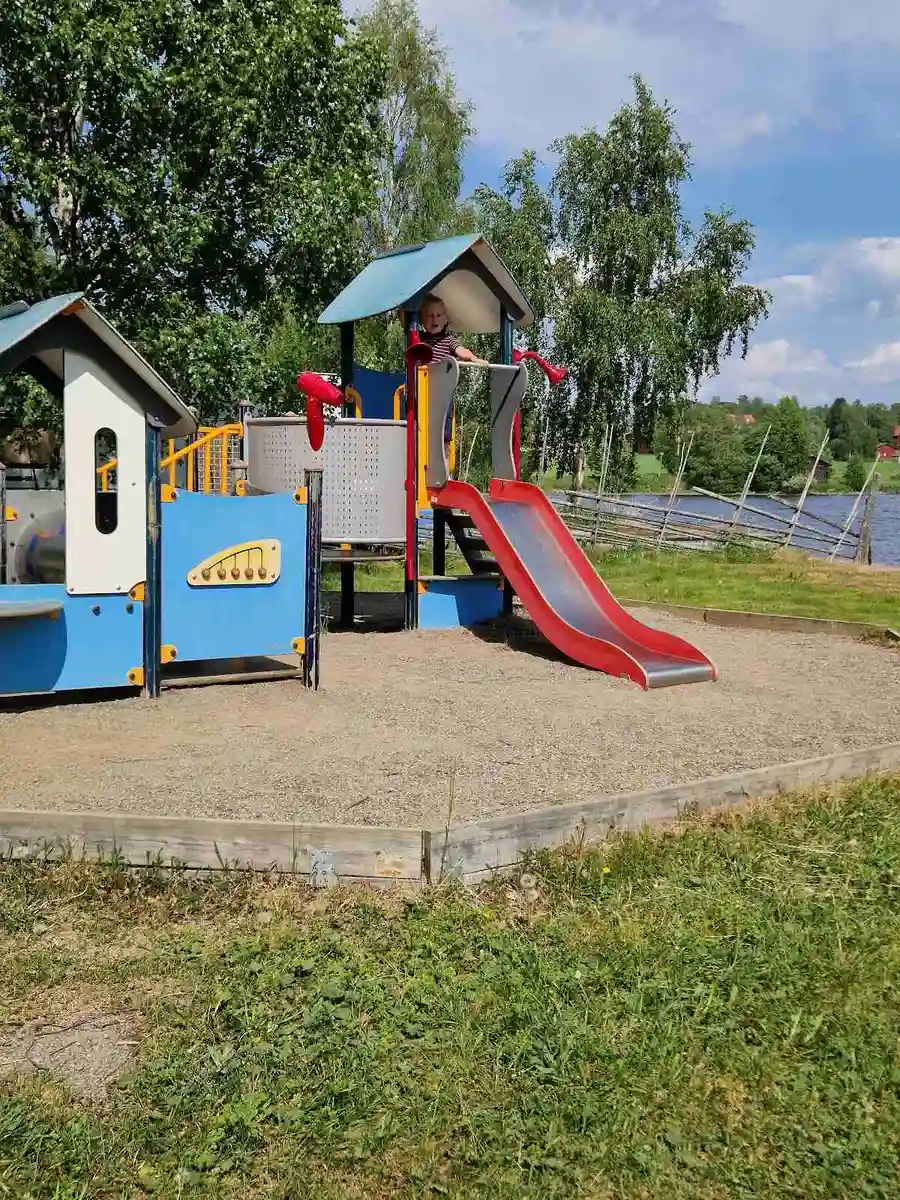 En lekplats på Hoverbergs camping med en rutschkana och annan utomhuslekutrustning i en parkmiljö, omgiven av grönska och viloplatser i skugga.