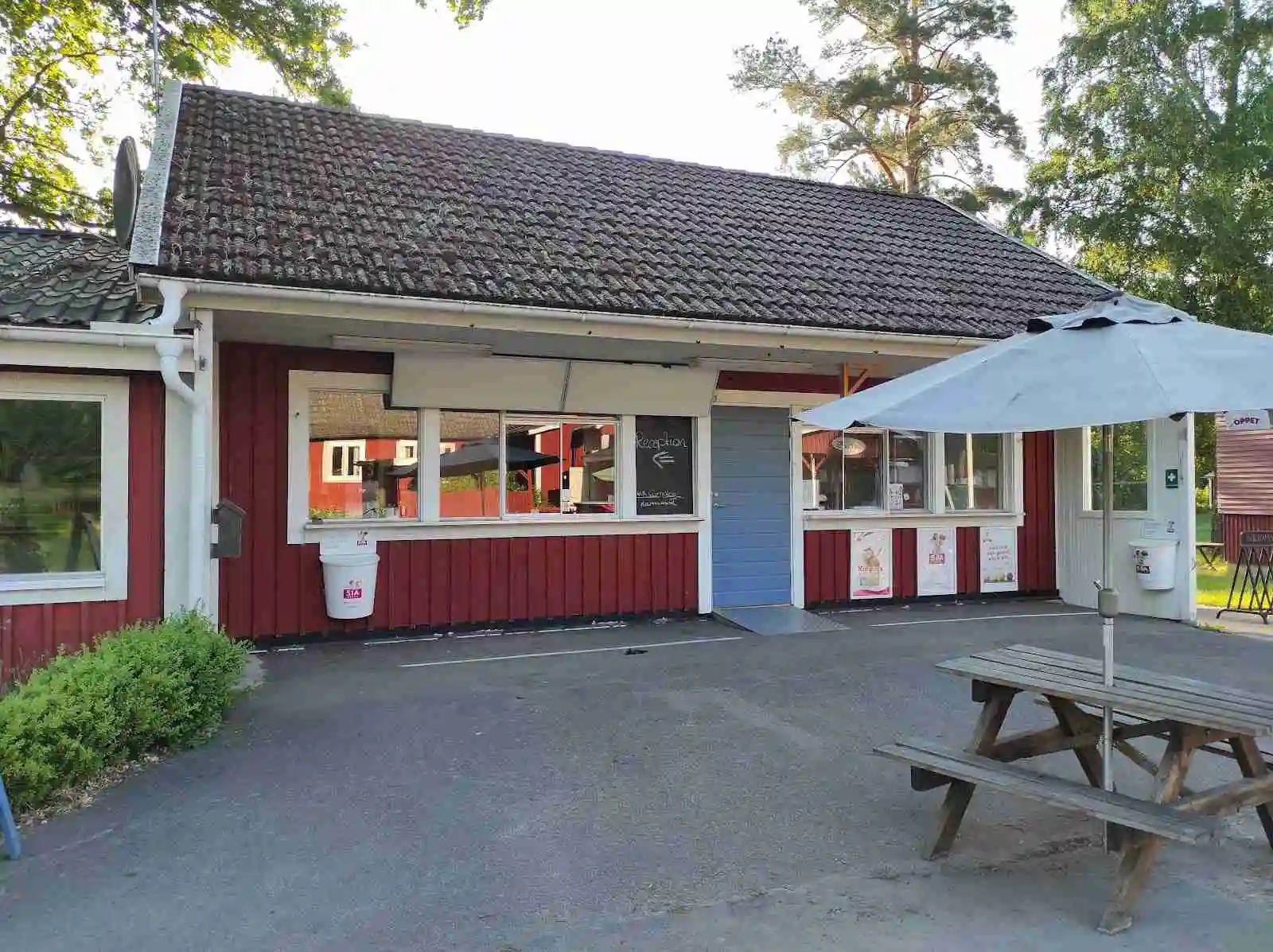 En mysig stuga med tak och träpanel, omgiven av grönskande natur på Hätteboda Vildmarkscamping. En grusad uppfart och vilområde syns framför stugan.