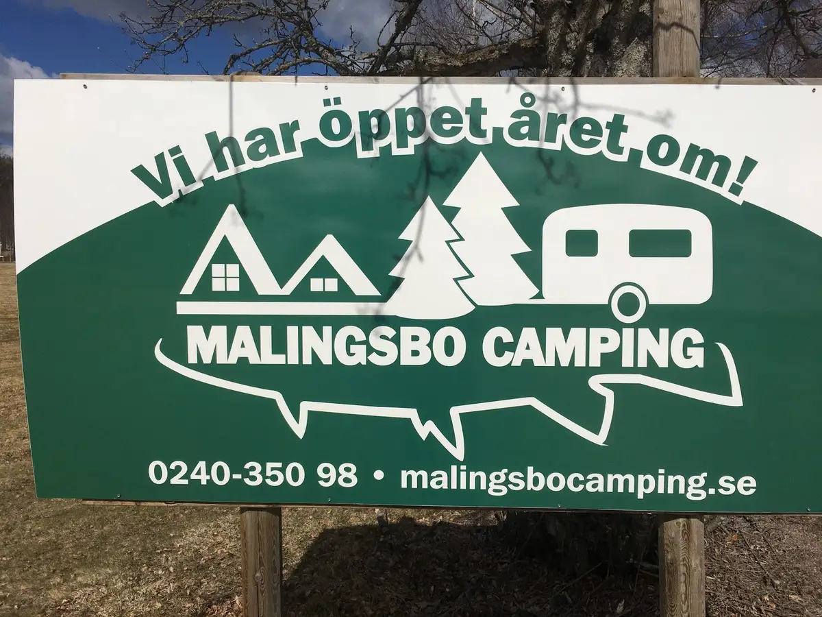 Bild av Malingsbo Camping: En campingplats med flera tält uppsatta i en naturskön miljö. En skylt med reklam och annan information syns i förgrunden. Det finns även tydliga trampade stigar som leder genom området.