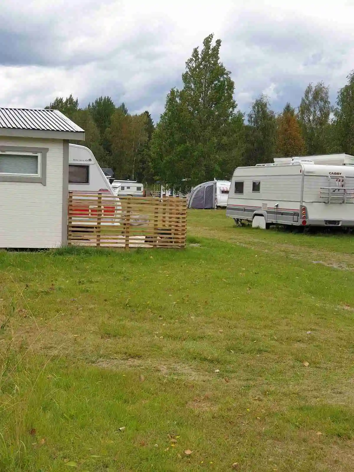 En campingplats med flera husvagnar och husbilar uppställda på en gräsmatta, omgiven av molntäckt himmel, träd och parkmiljö.