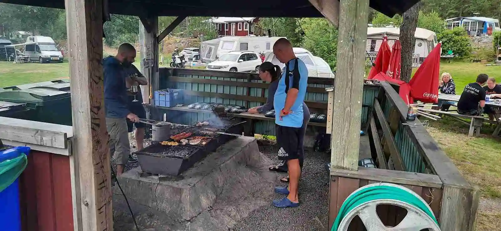 En familj som njuter av en måltid vid ett picknickbord vid Arkösunds camping, omgiven av grönska och friluftsmöbler.