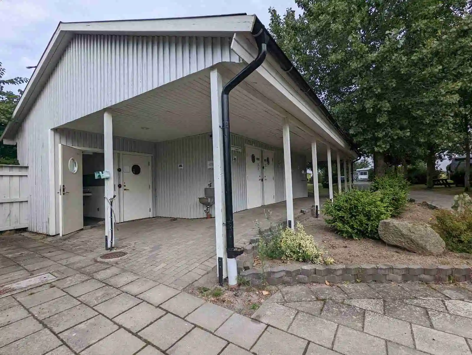 Bild av Fredriksbergs kursgård-camping som visar en bostadsområde med flera byggnader. Taken är tydligt synliga och omgivna av ett grönskande område. Asfalt är belagd på en uppfart framför byggnaderna, vilket kan indikera en entré eller väg in till platsen.