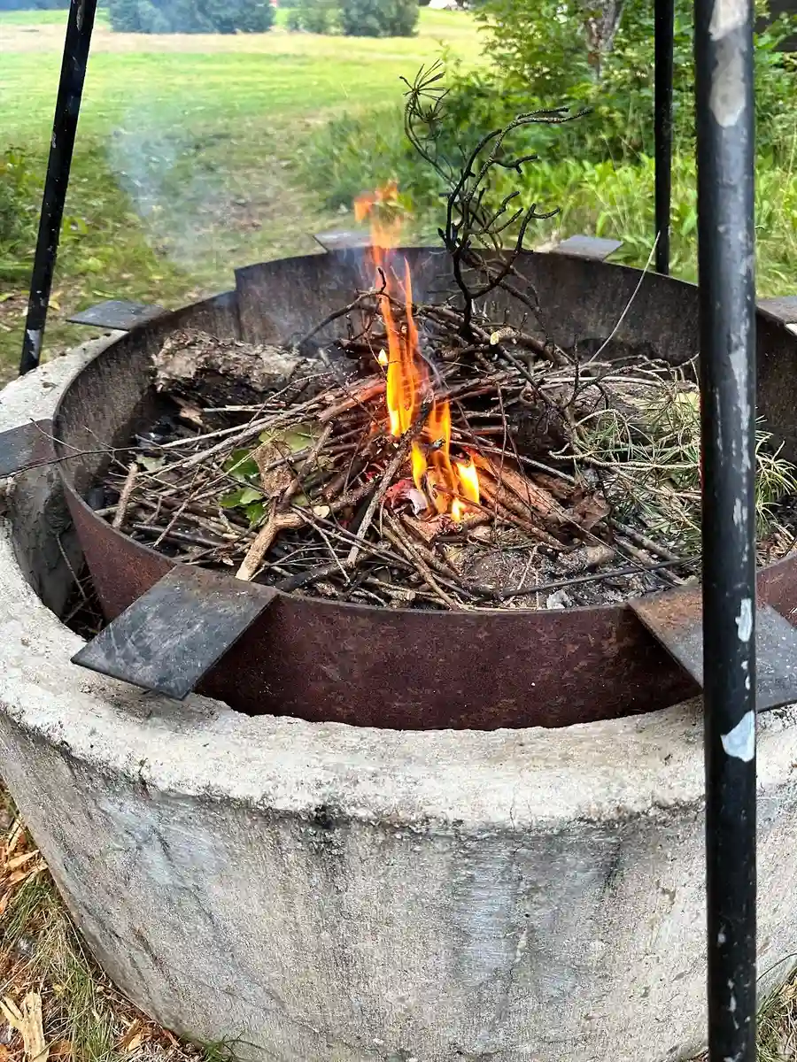 En brasa i vildmarken, omgiven av aska och glöd, med kokkärl uppvärmda över elden vid Hallsjö vildmarkscamping.