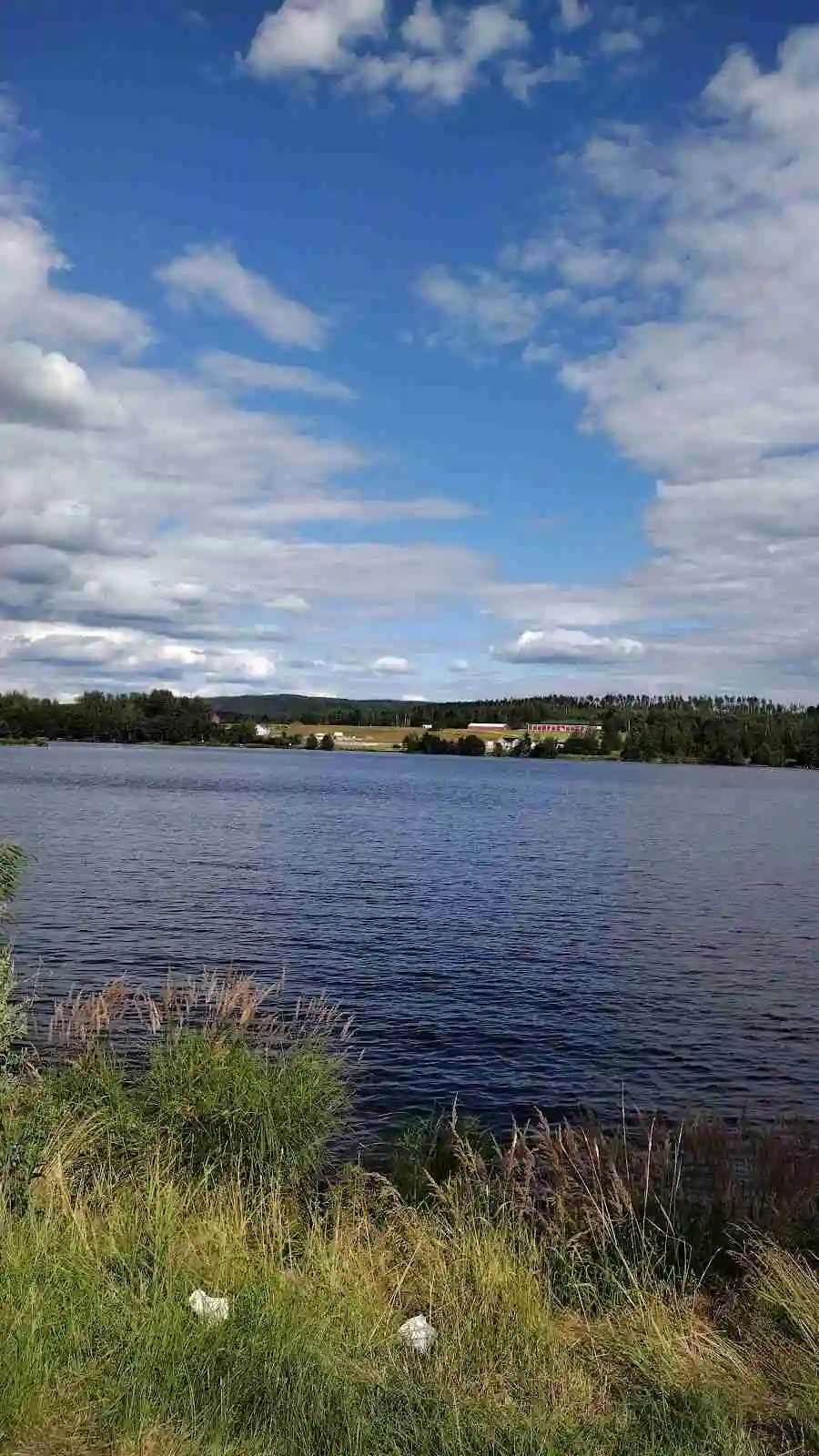 En bild av Sunne camping och sommarland vid en klar, blå himmel. Vattenområdet i förgrunden reflekterar molnen och skapar en lugn och naturskön atmosfär.
