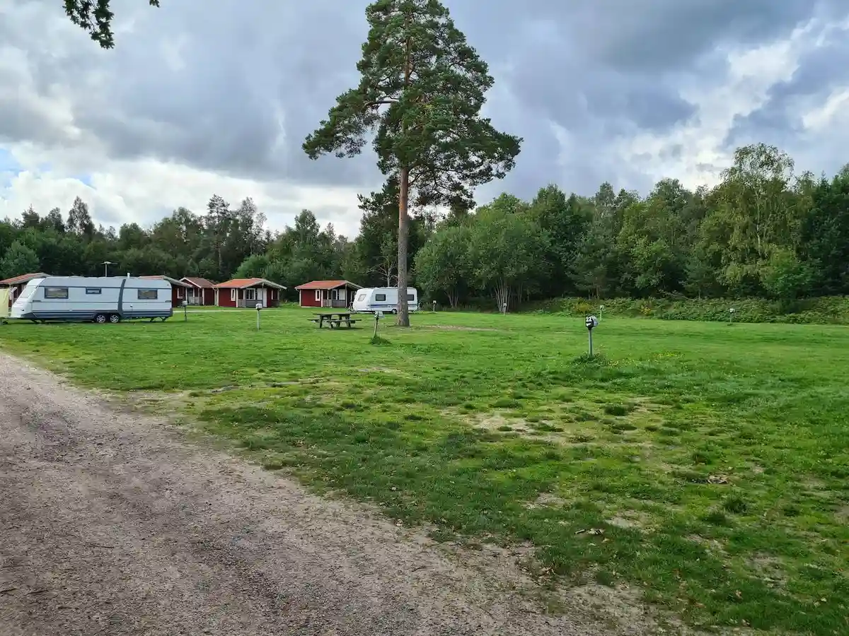 En grön campingplats med gräs och träd, moln på himlen och öppna ängar i bakgrunden.