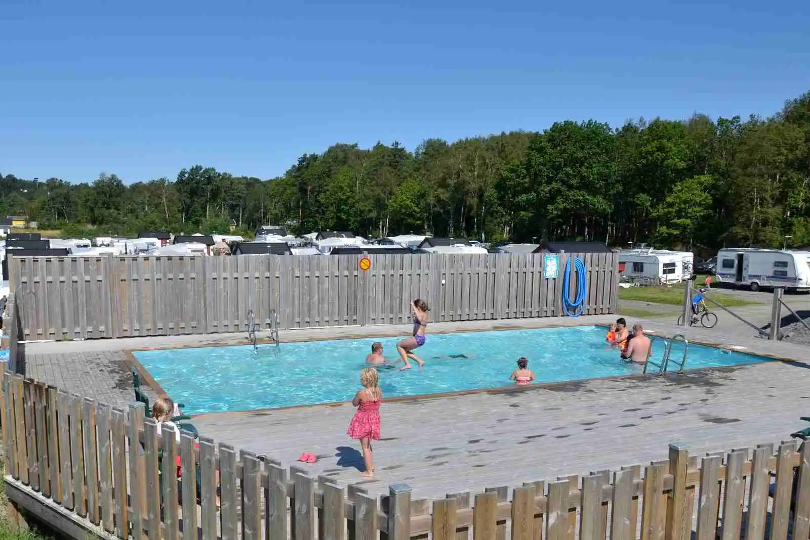 En solig dag på Stråvalla Camping med gäster som njuter vid poolen. Vuxna och barn i badkläder kopplar av och har roligt, omgivna av campingplatsens somriga atmosfär.