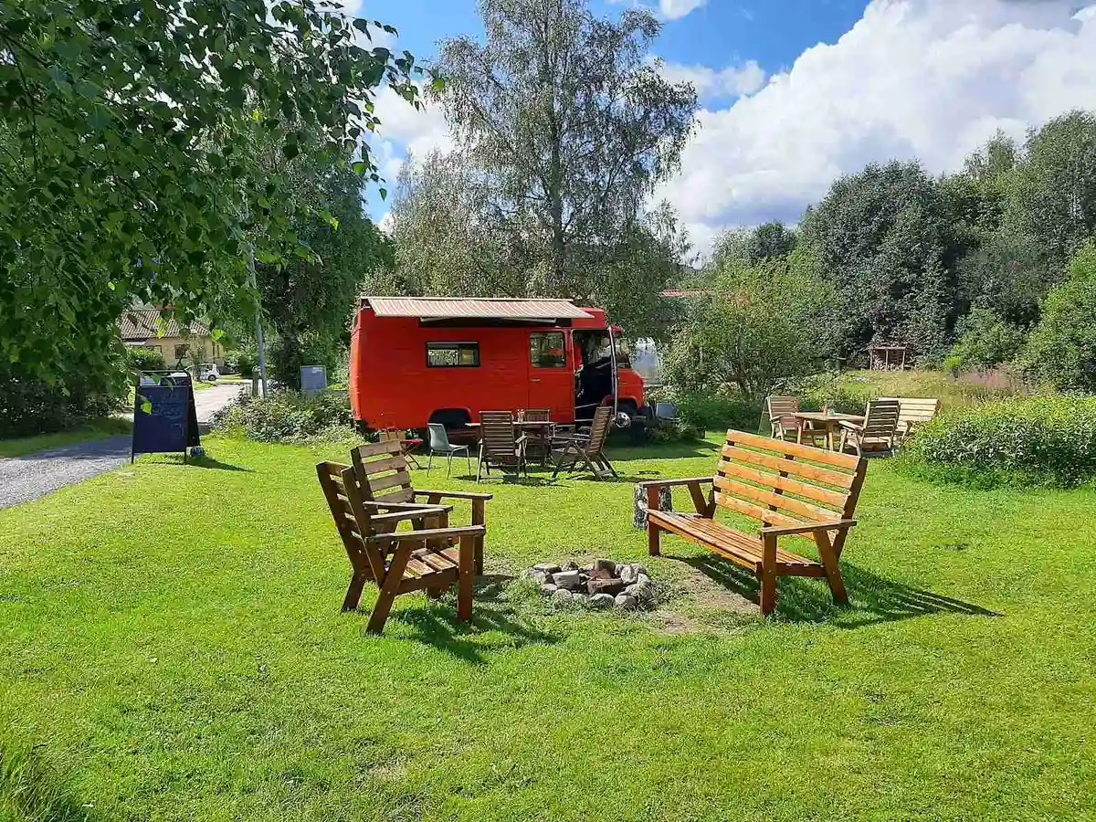 En grön campingplats med gräsmatta och trädgård, där en husvagn är parkerad. Området har utemöbler, inklusive ett bord, i vad som ser ut som en park eller bakgårdsmiljö.