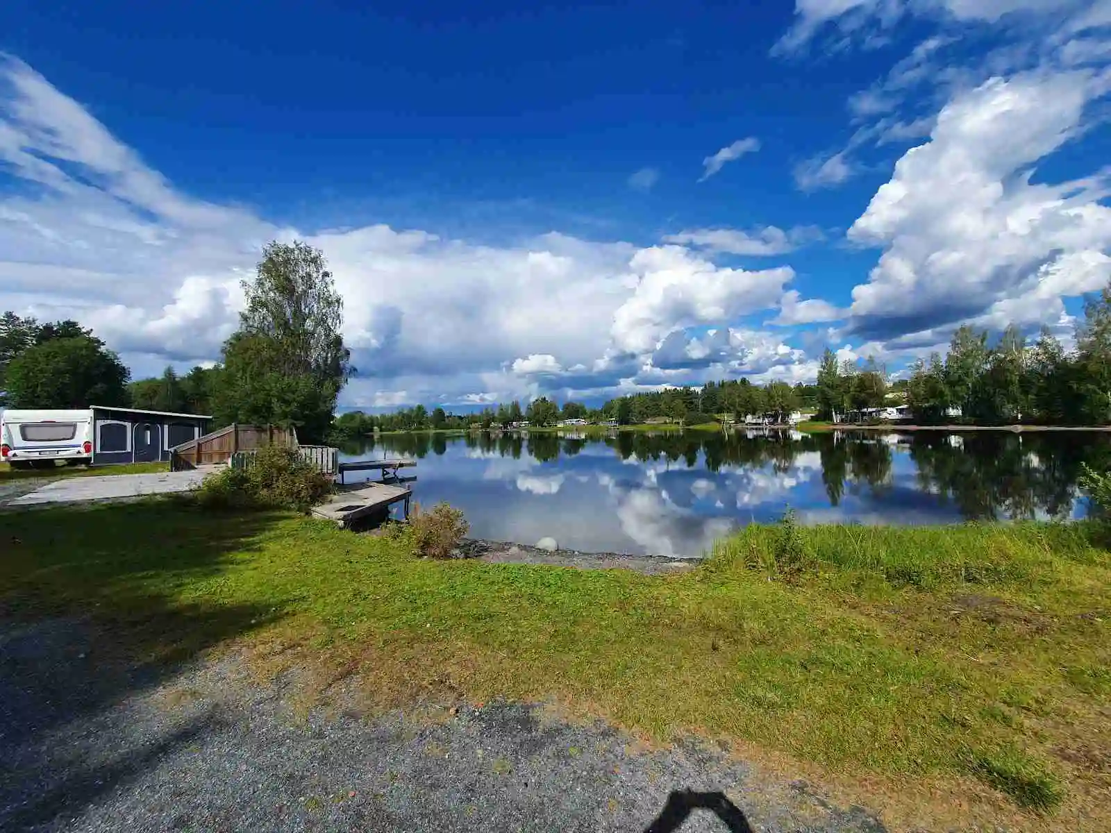 En naturskön vy över Krokomvikens camping, med en lugn sjö som speglar moln på himlen och omgivande gröna banker, som skapar en rofylld atmosfär.