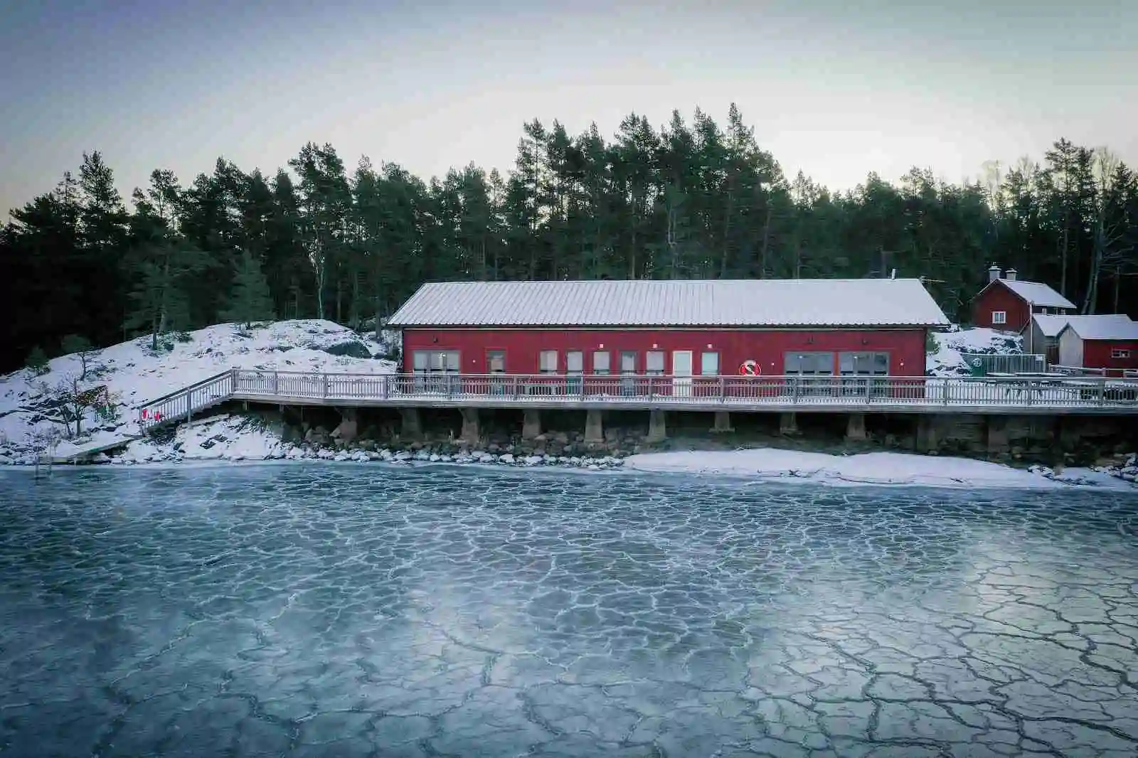 En vintrig kustvy vid Ängskärs hamncamping med snötäckt mark, isbelagda ytor och en liten timmerstuga. I bakgrunden syns en brygga och ett staket, allt täckt av ett lager snö. Reflektioner av vinterlandskapet kan skymtas i vattnet.