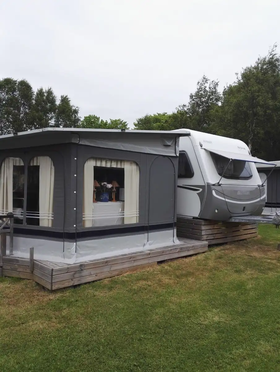 En campingplats med flera husvagnar och husbilar parkerade på gräsytan, omgiven av grönskande träd och klar himmel.