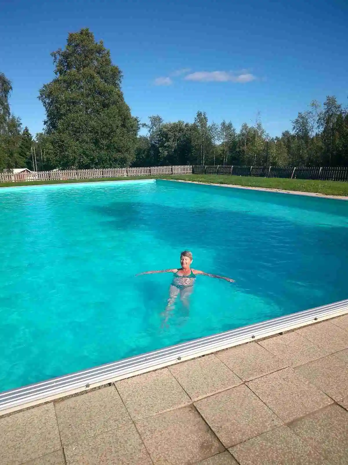 En utomhuspool med blått vatten omgiven av gröna träd, där gäster njuter av en avkopplande sommardag på Docksta Vandrarhem & Camping Kustladan.