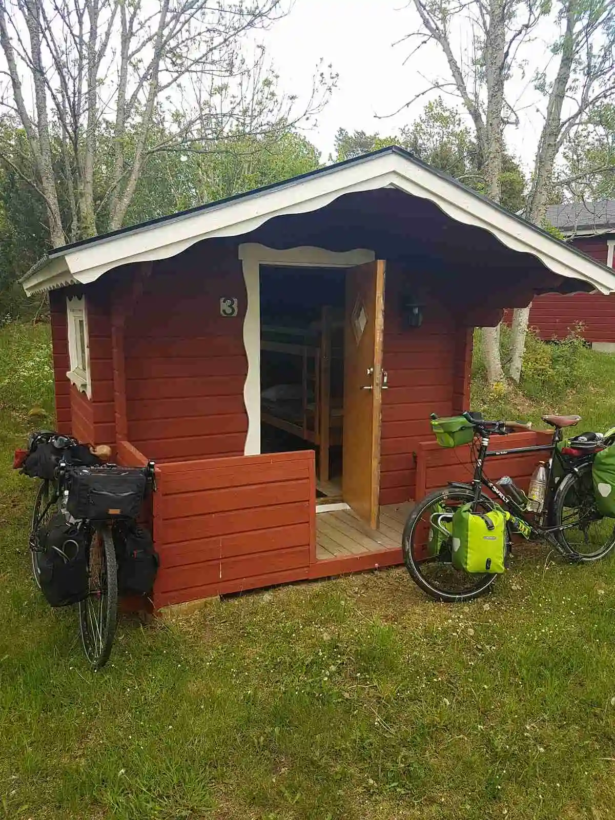 En trästuga med sidpaneler på en camping. Stugan är omgiven av grönska och ligger i en bakgårdsliknande miljö.