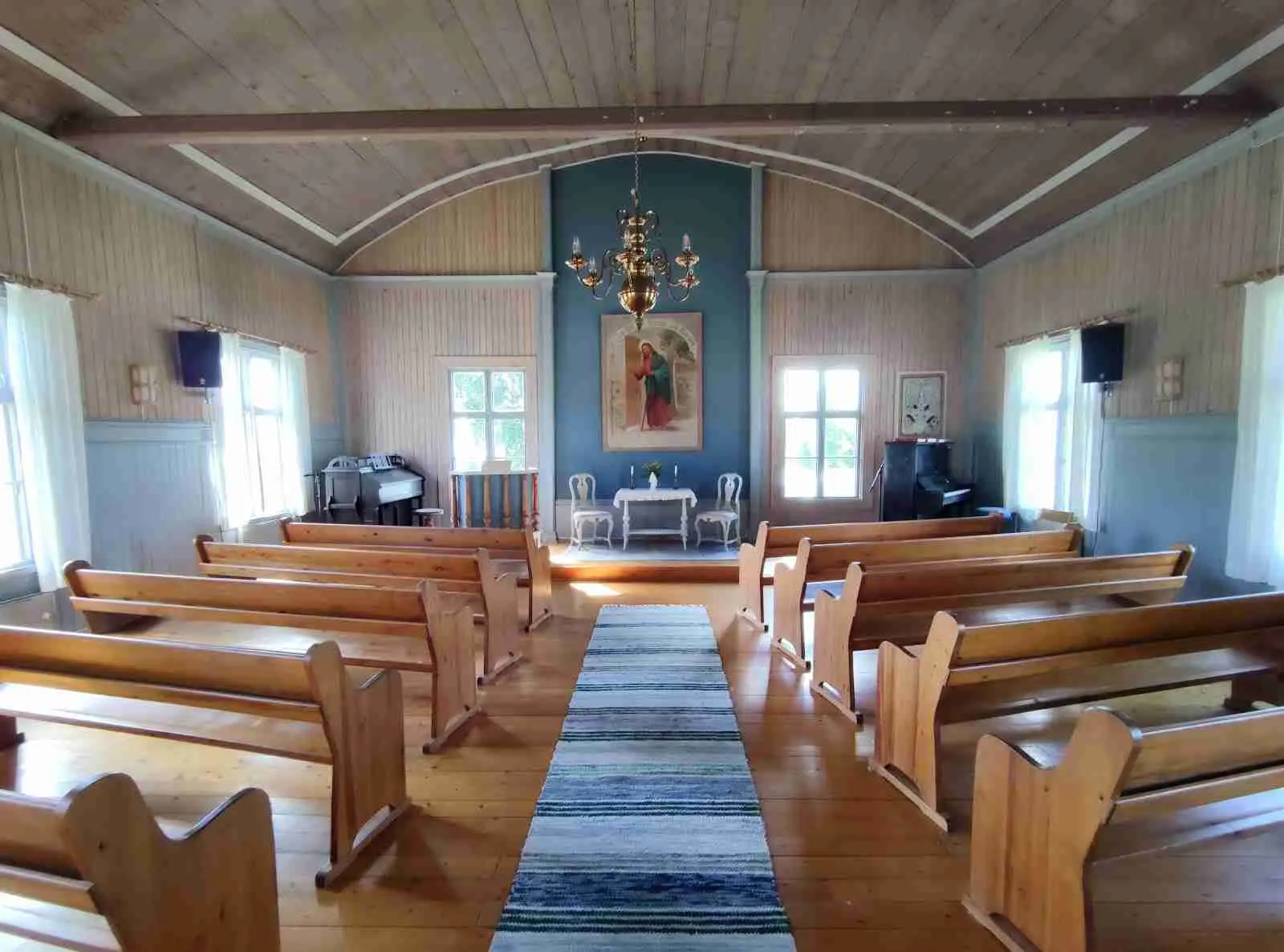 En ljus inomhusmiljö i Vikarbygården med trädetaljer och en rustik känsla. Ett välvt tak, väggpaneler och dörrar av trä ger intrycket av en gammaldags kyrksal eller kapell. Ljuskronor hänger från taket och en religiös dekoration pryder rummet.