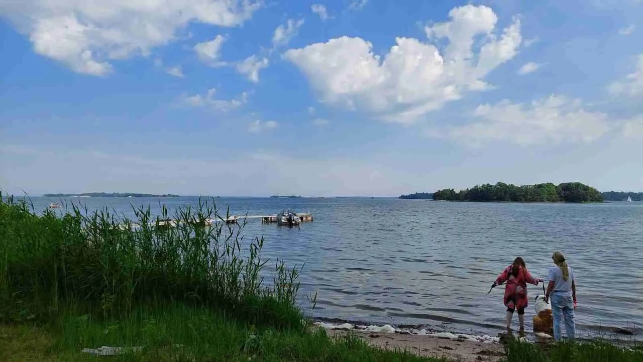 En vidsträckt vy över Kapellskärs camping med en klar himmel, sjö och kustlinje i bakgrunden. Marken och vattnet skapar en avkopplande atmosfär för rekreation vid sjön.