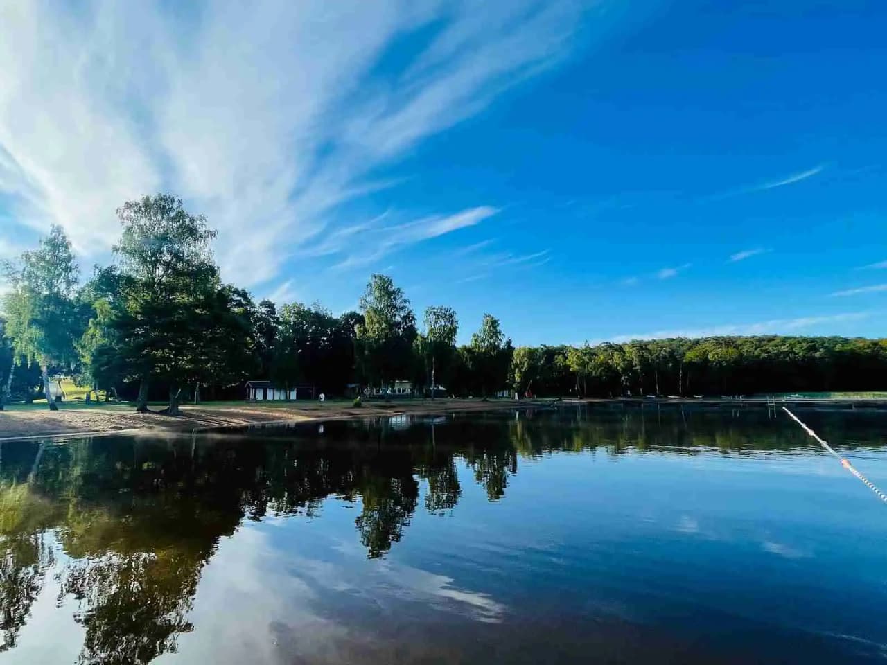 En lugn sjö med blått vatten omgiven av grönskande träd och natur, med spegelblanka reflektioner av den naturliga omgivningen vid Luhrsjöbadens camping.