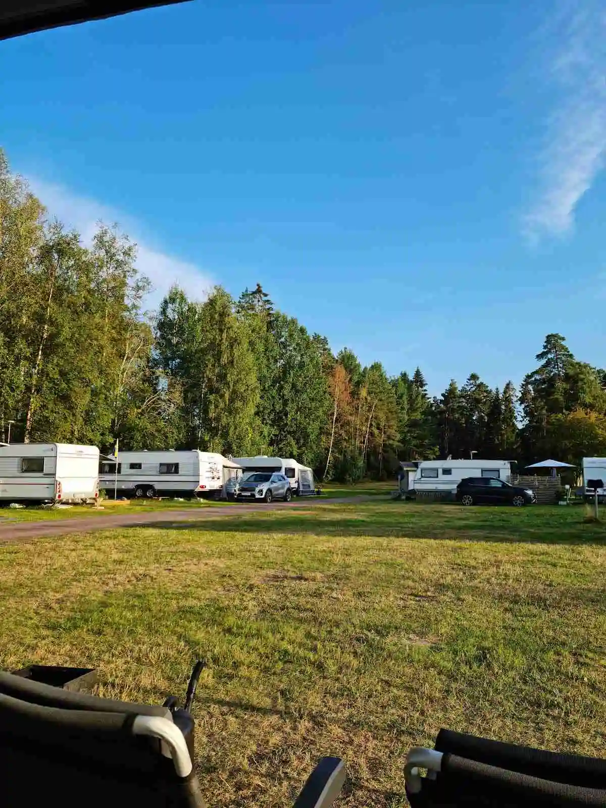En grön campingplats i Malexander med tält och husvagn uppställda på en gräsyta under en klar himmel. Området omges av träd som ger skugga. 