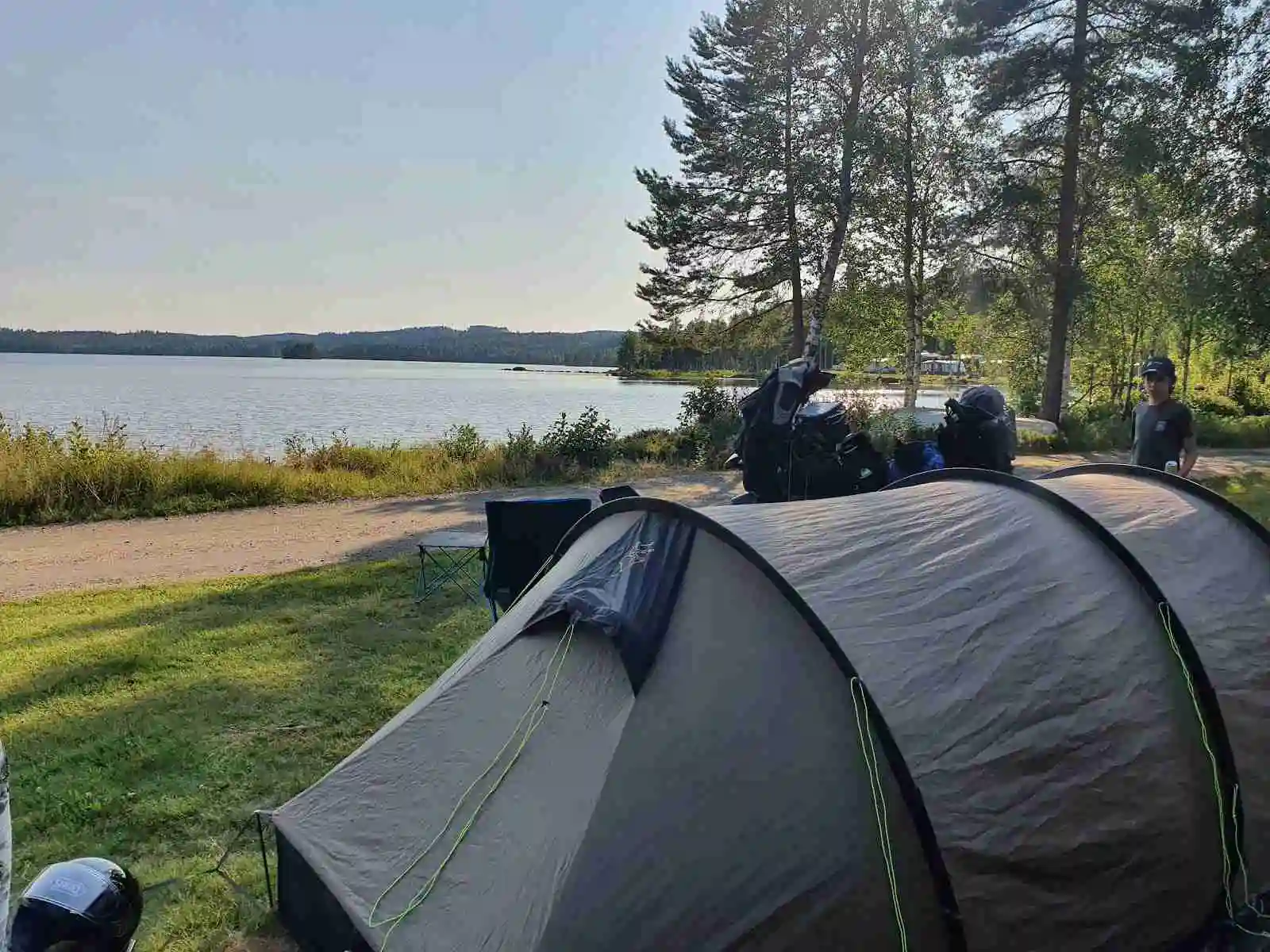 En bild av Treens Natur & Fiskecamp med tält uppsatta nära en sjö. Campingutrustning syns i förgrunden och en vacker landskap omger platsen, vilket skapar en avkopplande och naturnära atmosfär.